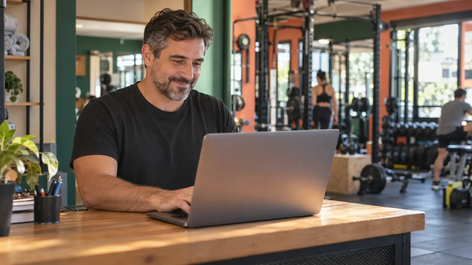 El dueño del gimnasio, en el mostrador, revisa su panel en la notebook con el gimnasio en marcha detrás.